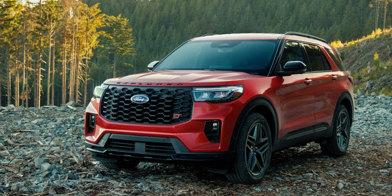 Front three-quarter view of a red 2026 Ford SUV parked on a rocky forest trail, featuring a black mesh grille, sporty accents, dark alloy wheels, and tall trees in the background under warm natural light.